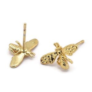 May include: Two gold-colored bee stud earrings. The earrings are detailed with wings, antennae, and body segments. The posts are visible on the back of the earrings. The earrings are on a white background.