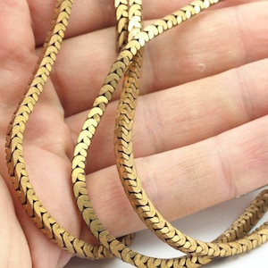 Könnte beinhalten: Eine goldene Schlangenkettenhalskette mit flachem, strukturiertem Design. Die Kette ist aus Metall und hat eine glänzende Oberfläche.
