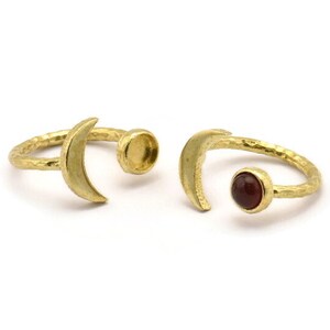 May include: Two gold crescent moon rings. One ring has a red gemstone set in the center, the other ring has a blank setting.