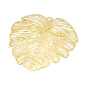 Può includere: Un ciondolo a forma di foglia in metallo traforato color oro, simile a una foglia di pianta Monstera. Il ciondolo ha un piccolo anello nella parte superiore per appenderlo ed è posto su uno sfondo bianco. Adatto per la creazione di gioielli o progetti artigianali.