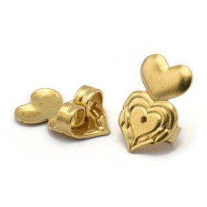 Könnte beinhalten: Zwei goldfarbene herzförmige Ohrstecker-Rückschlüsse. Ein Verschluss ist von vorne zu sehen, mit einem detaillierten Herzdesign. Der andere Verschluss ist von hinten zu sehen, mit einer glatten Herzform.