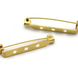 Könnte beinhalten: Zwei goldfarbene Metall-Sicherheitsnadeln mit jeweils vier Löchern. Die Sicherheitsnadeln sind zum Befestigen von Broschen, Anstecknadeln oder anderem Schmuck an Kleidung gedacht.