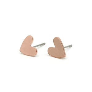 Op de afbeelding: Een paar hartvormige oorknopjes in roségoud. Elke oorknop heeft een glad, plat hartontwerp met een metallic afwerking en een dunne, zilverkleurige stift aan de achterkant, ontworpen voor gaatjes in de oren en een stevige bevestiging.