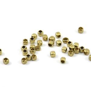 Puede incluir: Numerosas cuentas pequeñas, doradas y con forma de cubo están dispersas sobre una superficie blanca. Cada cuenta tiene un agujero en el centro. Las cuentas están dispersas, algunas más juntas y otras más aisladas.