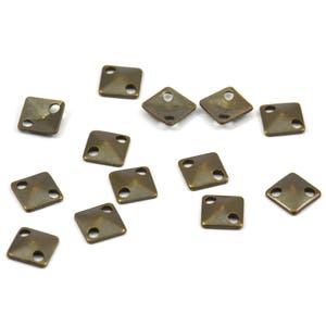 May include: A collection of 14 antique gold-toned metal square-shaped embellishments with two holes each. The embellishments are scattered on a white surface.