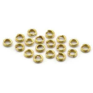 Op de afbeelding: Een groep van 18 gouden springringen, ook wel splitringen genoemd, liggen verspreid op een wit oppervlak. Deze ringen worden gebruikt om juweelcomponenten te verbinden.