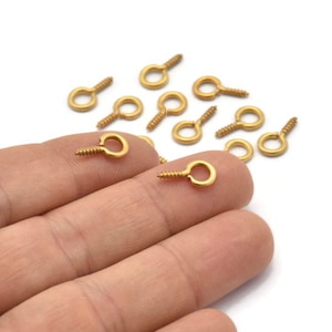 May include: A close-up of a hand holding a group of gold-toned screw-in eyelet hooks. The hooks are small and have a round eyelet with a screw thread.