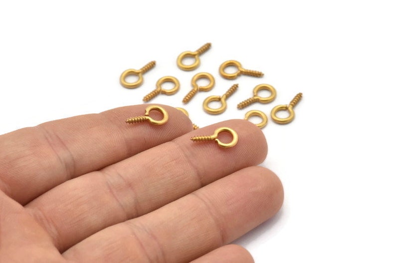 May include: A close-up of a hand holding a group of gold-toned screw-in eyelet hooks. The hooks are small and have a round eyelet with a screw thread.