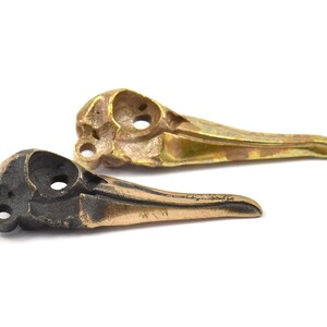 Op de afbeelding: Twee vogel schedel replica's, &eacute;&eacute;n in brons en &eacute;&eacute;n in goud, tegen een witte achtergrond. De schedels hebben oogkassen en snavelvormen, wat een gotisch of macaber thema suggereert. De afbeelding toont de gedetailleerde afwerking.