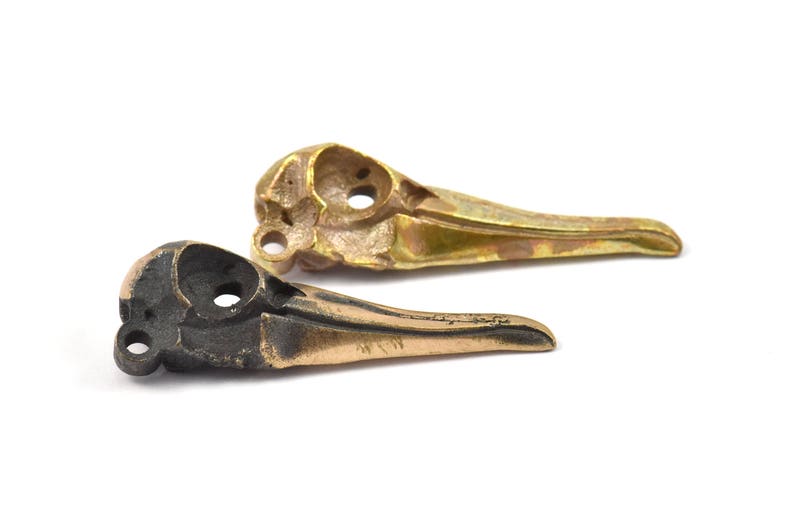 Op de afbeelding: Twee vogel schedel replica's, &eacute;&eacute;n in brons en &eacute;&eacute;n in goud, tegen een witte achtergrond. De schedels hebben oogkassen en snavelvormen, wat een gotisch of macaber thema suggereert. De afbeelding toont de gedetailleerde afwerking.