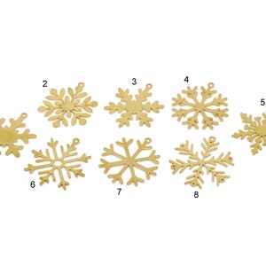 May include: Eight gold-coloured snowflake ornaments in various designs. Each snowflake has a small hole at the top for hanging. The ornaments are displayed against a white background, suitable for festive decorations.