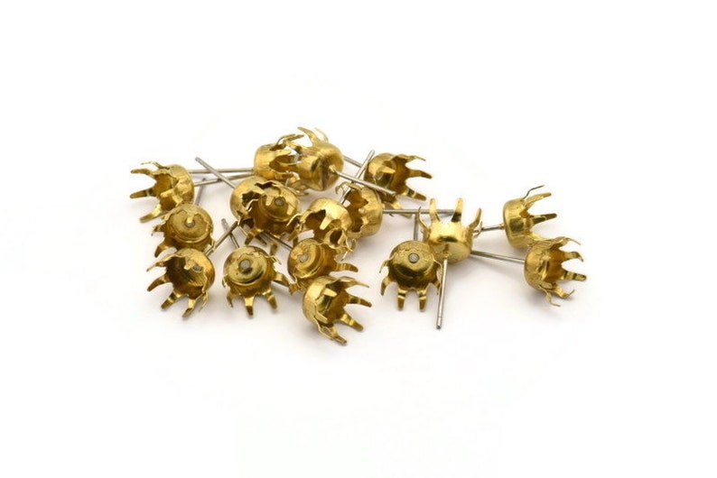 May include: A group of gold-coloured metal earring settings with a pointed end and a crown-shaped top. The settings are scattered on a white surface.