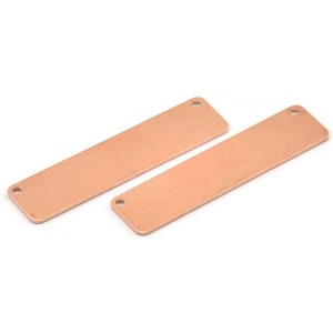 May include: Two rectangular, copper-colored metal blanks with rounded corners and two small holes. The blanks are lying on a white surface. These blanks are suitable for jewelry making or crafting.