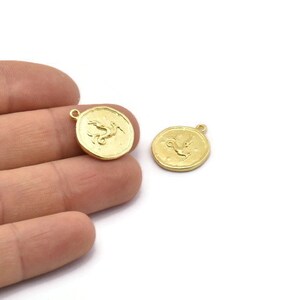 Op de afbeelding: Twee goudkleurige metalen bedels in de vorm van een ronde munt met een reli&euml;fdesign van een mythisch wezen. De bedels hebben een gat aan de bovenkant om aan een ketting of armband te bevestigen.