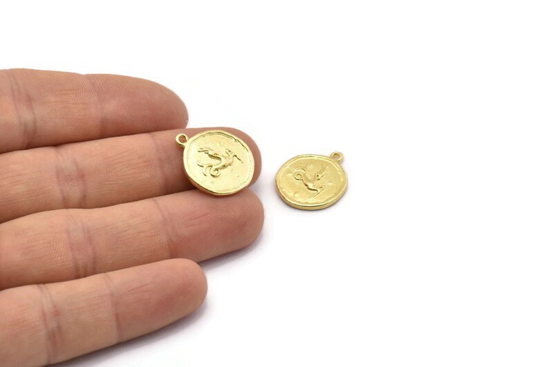 Op de afbeelding: Twee goudkleurige metalen bedels in de vorm van een ronde munt met een reli&euml;fdesign van een mythisch wezen. De bedels hebben een gat aan de bovenkant om aan een ketting of armband te bevestigen.