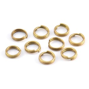 Puede incluir: Diez anillos de salto de metal de color dorado, cada uno con un diseño de doble bucle. Los anillos están dispuestos en un grupo suelto sobre un fondo blanco.