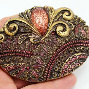 May include: A gold and red ornate oval brooch with a large, glittery, orange-red stone in the center.