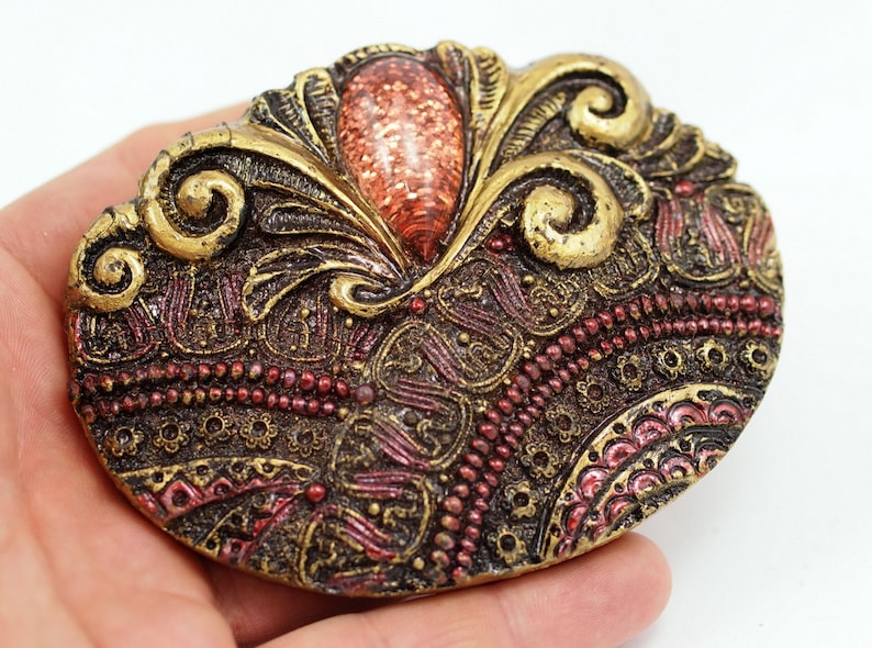 May include: A gold and red ornate oval brooch with a large, glittery, orange-red stone in the center.