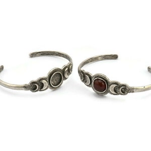 以下が含まれることがあります： 2つのシルバーカラーのカフブレスレット。各ブレスレットは、三日月形に挟まれた、小さな丸い石または空のスペースを備えた楕円形の中央デザインが特徴です。 ブレスレットはテクスチャ加工されたアンティーク仕上げで、白い背景に表示されています。