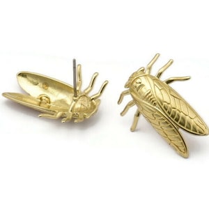 May include: A pair of gold-tone cicada earrings. The earrings are made of metal and have a detailed design. The cicadas are facing forward with their wings spread out.