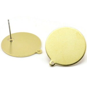 Puede incluir: Dos blancos de pendientes redondos de color dorado con un poste en un lado y un anillo de salto en el otro. Los blancos de pendientes son lisos y tienen una superficie ligeramente texturizada.