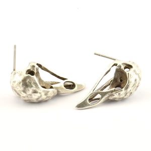 Puede incluir: Un par de pendientes de plata con forma de cráneos de pájaro. Los pendientes son detallados y tienen un acabado texturizado.