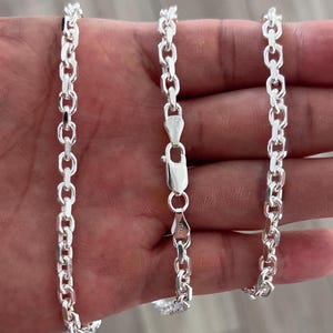 Puede incluir: Un collar de cadena plateada con un diseño de eslabones ovalados. El collar se muestra sobre un fondo neutro. La cadena está hecha de un metal brillante y reflectante y tiene un cierre de cierre.