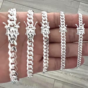 May include: A collection of silver-colored curb chain bracelets in varying widths. Each bracelet features a secure clasp. The bracelets are displayed on a hand, showcasing the different sizes available. The chains have a shiny, reflective surface.