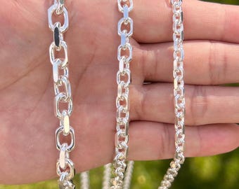 May include: Three silver chains of varying thicknesses are displayed side by side. The leftmost chain features large, elongated oval links, while the middle and right chains consist of smaller, uniform round links with a polished, reflective finish.