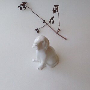 May include: A white ceramic figurine of a dog sitting with its head tilted up. The dog has a simple, realistic design.