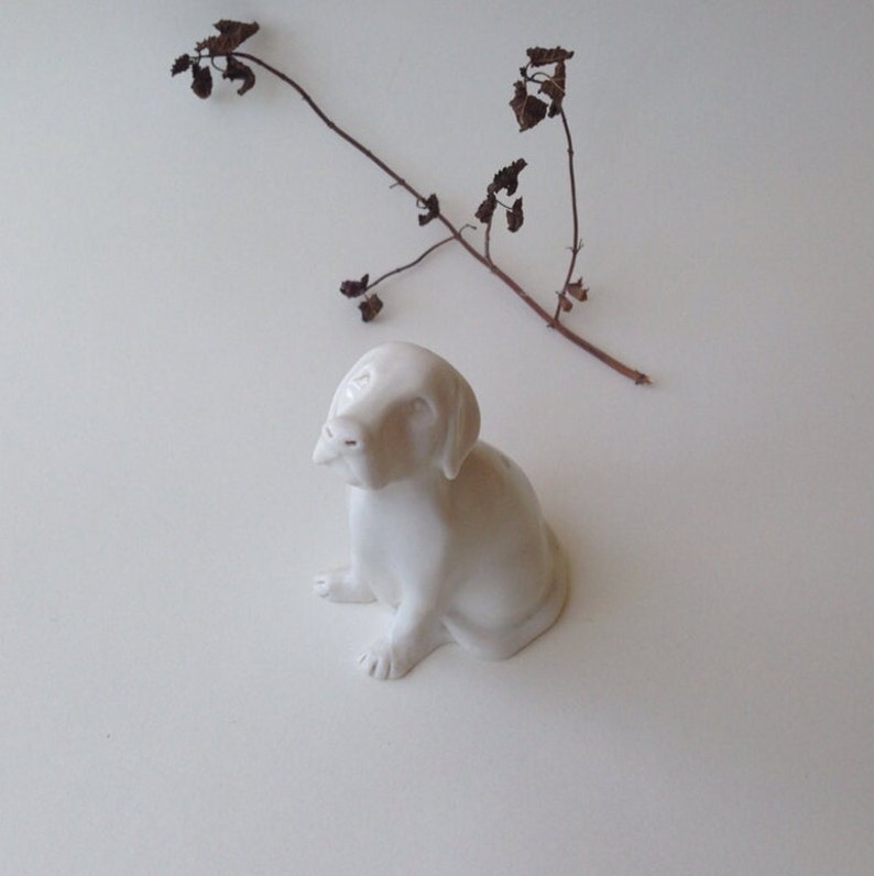 May include: A white ceramic figurine of a dog sitting with its head tilted up. The dog has a simple, realistic design.