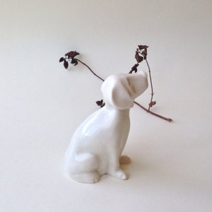May include: A white ceramic figurine of a dog sitting with its head tilted up. The dog is made of smooth, white porcelain and has a simple, elegant design.