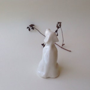 May include: White ceramic figurine of a dog sitting with its head tilted up. The figurine is on a white background.