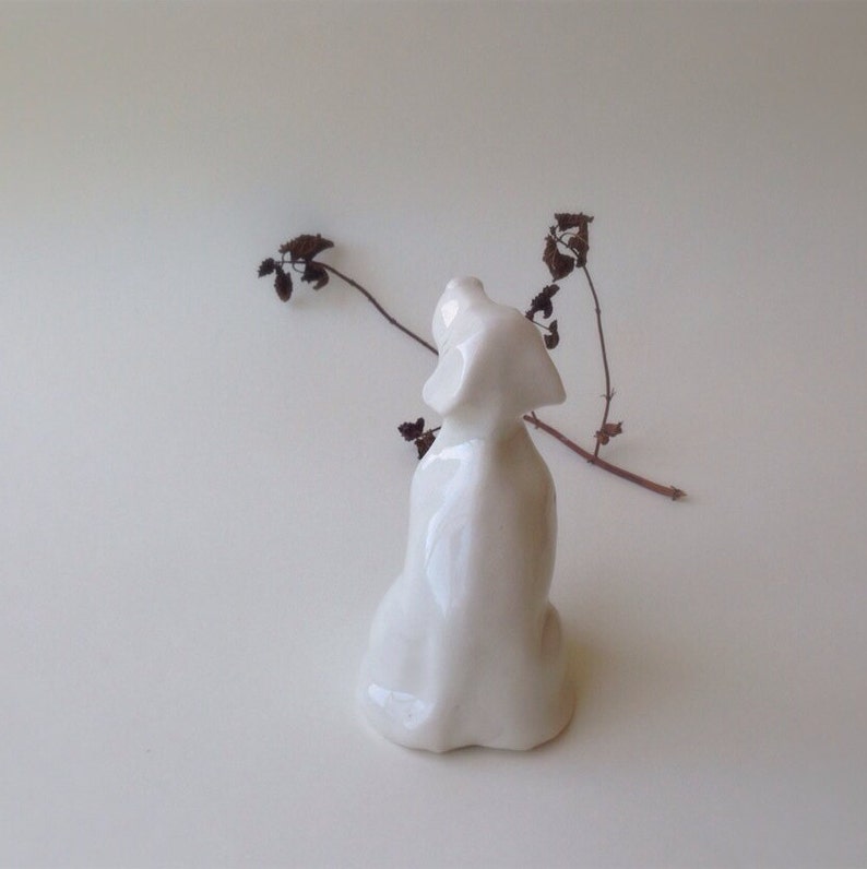 May include: White ceramic figurine of a dog sitting with its head tilted up. The figurine is on a white background.