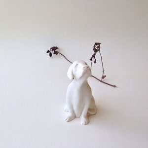 May include: White ceramic figurine of a dog sitting with its head tilted up, looking to the side. The dog is on a white background with a dried branch in the background.