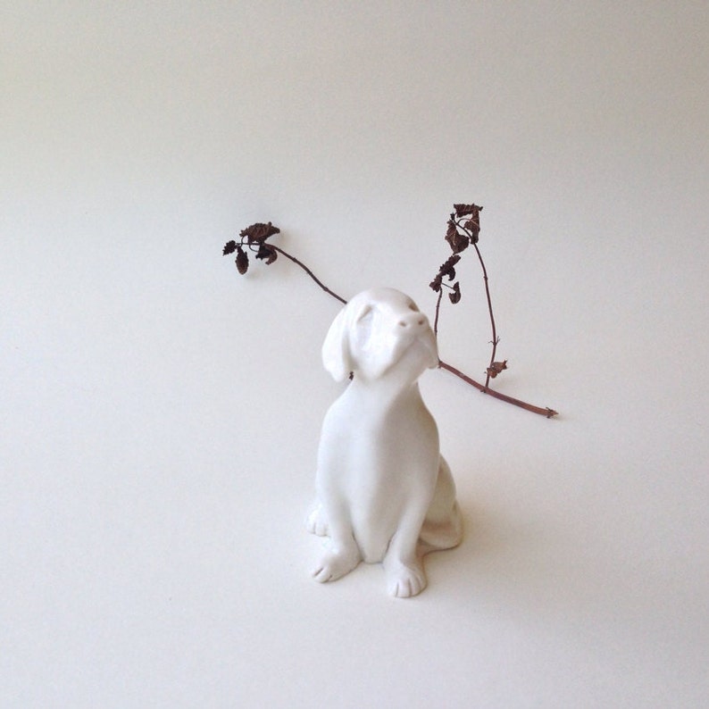 May include: White ceramic figurine of a dog sitting with its head tilted up, looking to the side. The dog is on a white background with a dried branch in the background.