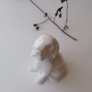 May include: A white ceramic figurine of a dog sitting with its head tilted up. The dog has a simple, stylized design.
