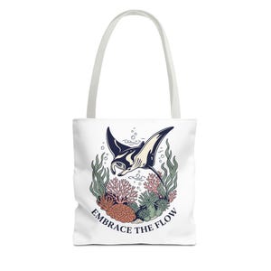 May include: White tote bag featuring a manta ray illustration in navy blue and cream, surrounded by coral and seaweed. The text "EMBRACE THE FLOW" is printed below the illustration. The bag has white handles.