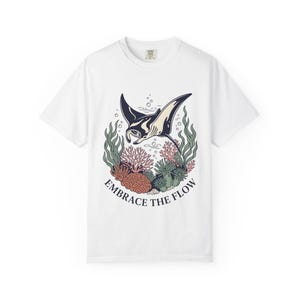 Op de afbeelding: Wit T-shirt met een afbeelding van een manta rog die boven koraal en zeewier zwemt. Het ontwerp bevat de tekst "Embrace the Flow". De manta rog is zwart en wit, en het koraal is rood en bruin.