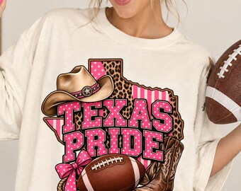 May include: An off-white t-shirt featuring a graphic of the state of Texas with leopard print and pink stripes. The design includes a cowboy hat, a brown football, a cowboy boot, a pink polka-dot bow, and the text Texas Pride in pink letters.