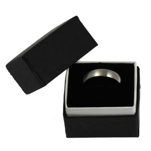 Product image of a 5mm brushed stainless steel band ring displayed inside an open black jewelry gift box. The box has a matte black exterior with a white inner border and a soft black velvet insert holding the ring upright. The ring features a matte silver tone brushed finish with a smooth, minimalist design and rounded edges, presented against a plain white background.