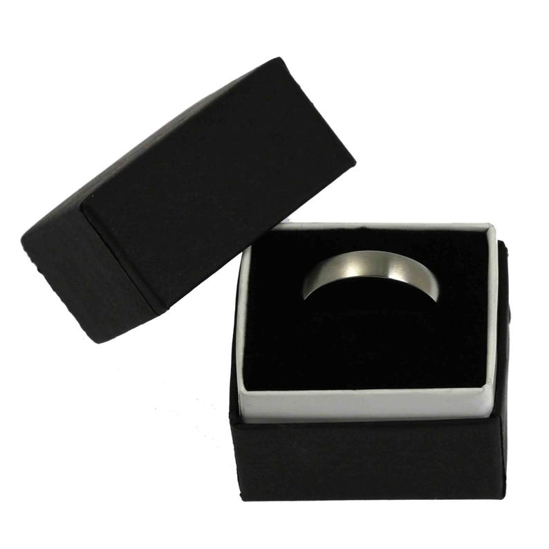 Product image of a 5mm brushed stainless steel band ring displayed inside an open black jewelry gift box. The box has a matte black exterior with a white inner border and a soft black velvet insert holding the ring upright. The ring features a matte silver tone brushed finish with a smooth, minimalist design and rounded edges, presented against a plain white background.