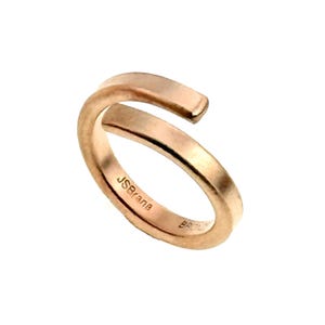 May include: A rose gold-toned bypass ring with a smooth, polished finish. The interior of the band features the engraved text JSBrana and additional markings. The ring has an open design with two overlapping ends, creating a continuous circular shape.