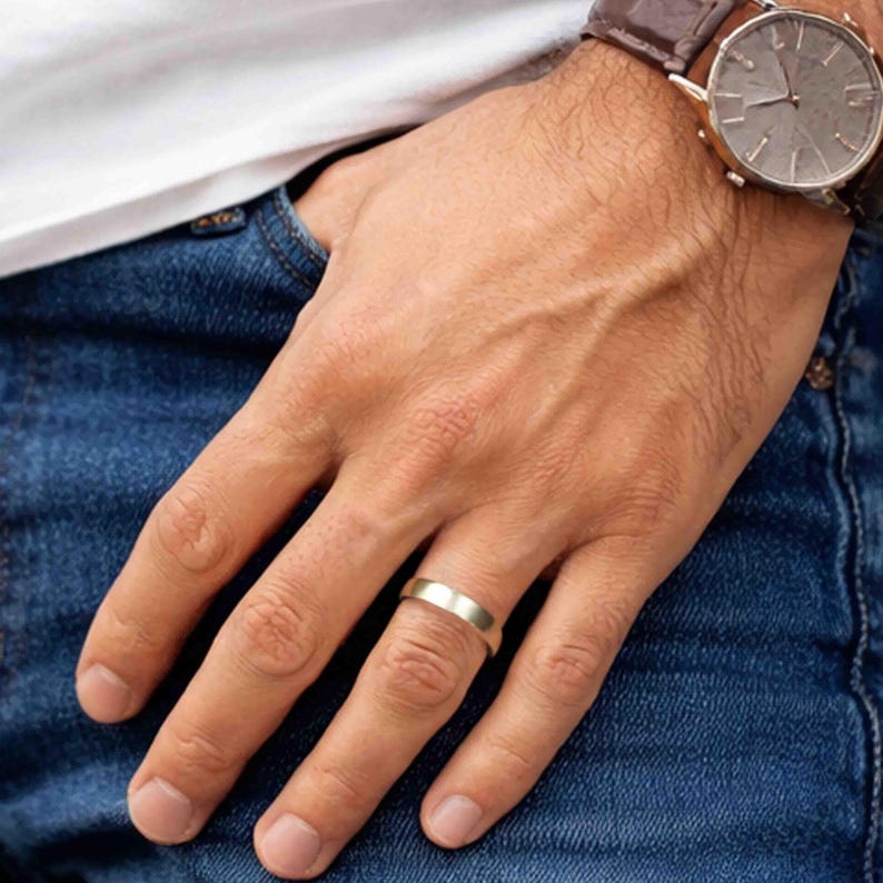 Close-up lifestyle image of a hand resting against blue denim jeans, wearing a 5mm brushed stainless steel band ring on the ring finger. The ring has a matte silver tone finish with a smooth, minimalist design and softly rounded edges. The person is also wearing a brown leather strap wristwatch with a silver case. The clean, modern band complements a casual everyday outfit.