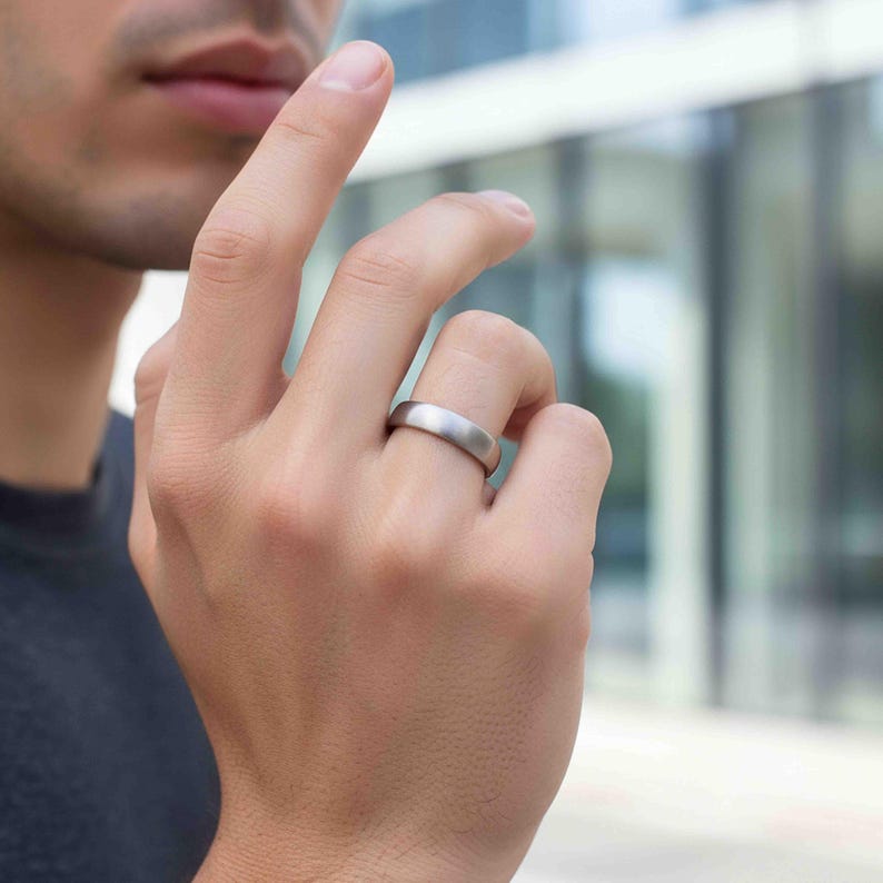 Close-up lifestyle image of a man wearing a 5mm brushed stainless steel band ring on their ring finger. The matte silver tone ring has a smooth, minimalist design with a soft brushed finish. The hand is raised near the face, and the background shows a blurred modern outdoor setting with glass and metal elements. The unisex wedding band features a clean, sleek profile suitable for everyday wear.