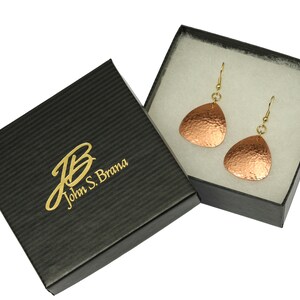 May include: A pair of copper earrings with a hammered texture, hanging from gold hooks, in a black gift box with the text "John S. Brama" in gold lettering.