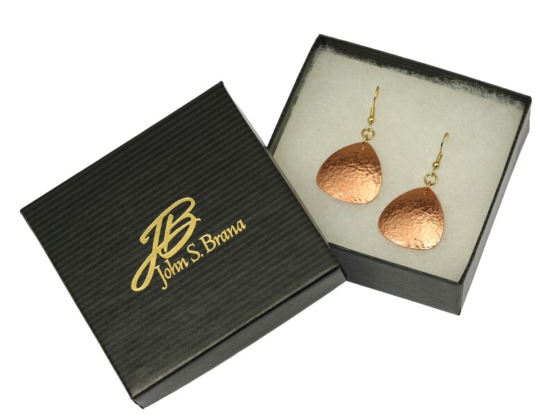 May include: A pair of copper earrings with a hammered texture, hanging from gold hooks, in a black gift box with the text "John S. Brama" in gold lettering.