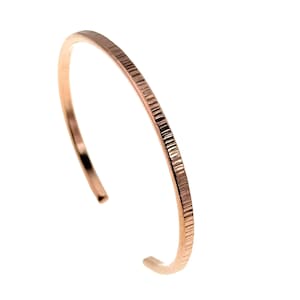 May include: A rose gold cuff bracelet with a textured, linear pattern. The open design allows for easy wear. The bracelet is a simple, elegant piece of jewellery.