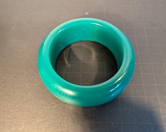 May include: A teal bangle bracelet. The bracelet is a solid, smooth band with a rounded shape. The colour is a vibrant, glossy teal, and the bracelet appears to be made of a solid material. The bangle is a simple, yet bold piece of jewellery.