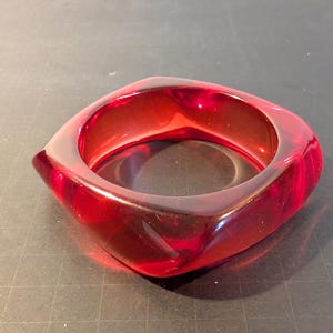 May include: A square-shaped, translucent red bangle bracelet. The bracelet is made of a glossy material and has rounded corners. The light reflects off the surface, creating highlights and shadows. The bracelet is a bold accessory.